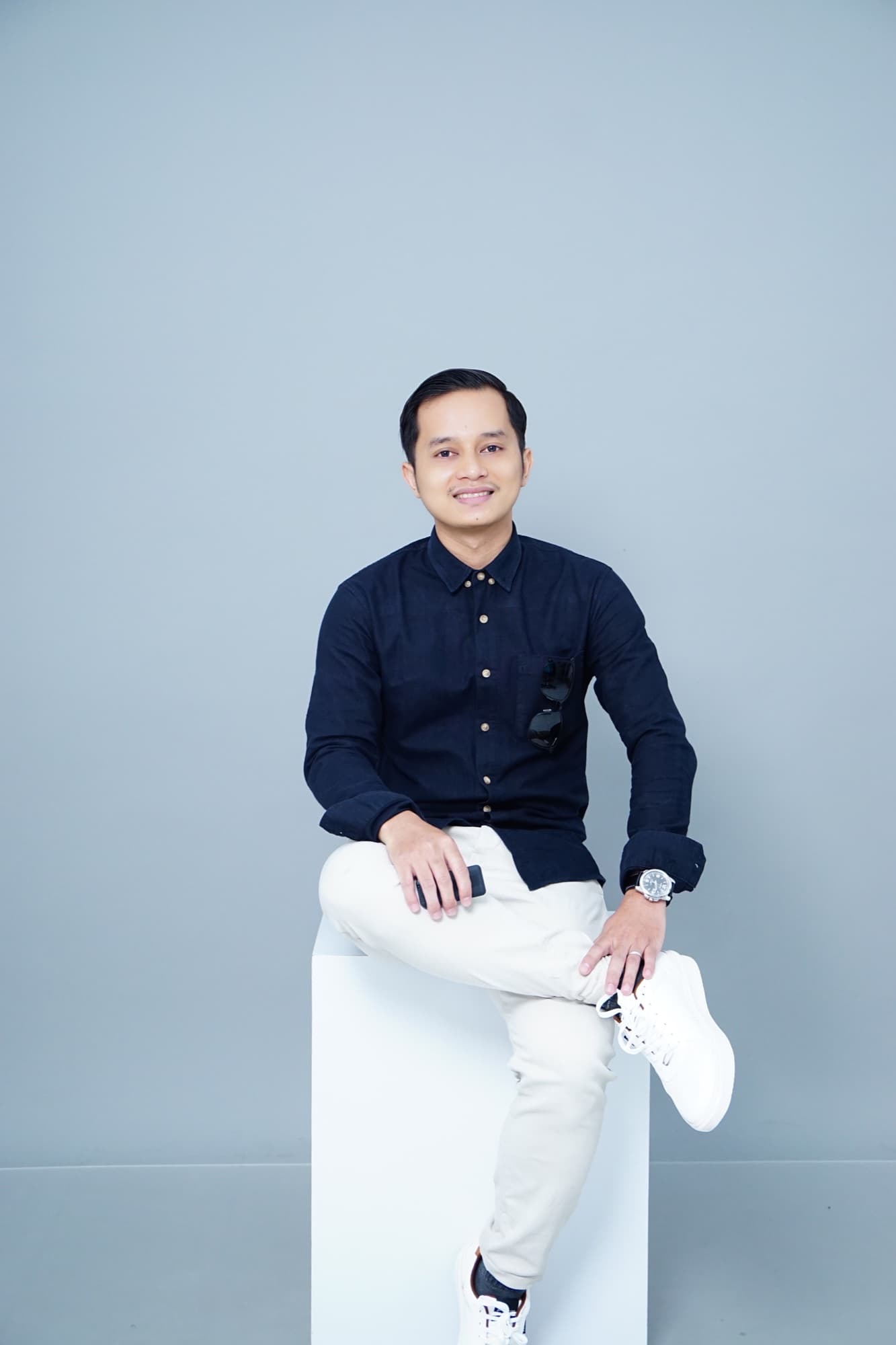 Hilmi sitting on a studio stool, wearing a navy shirt and white trousers